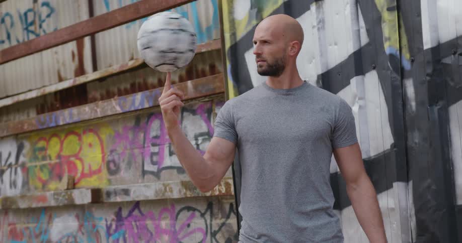 Man spins a soccer ball on his finger while standing in front of a graffiti covered building