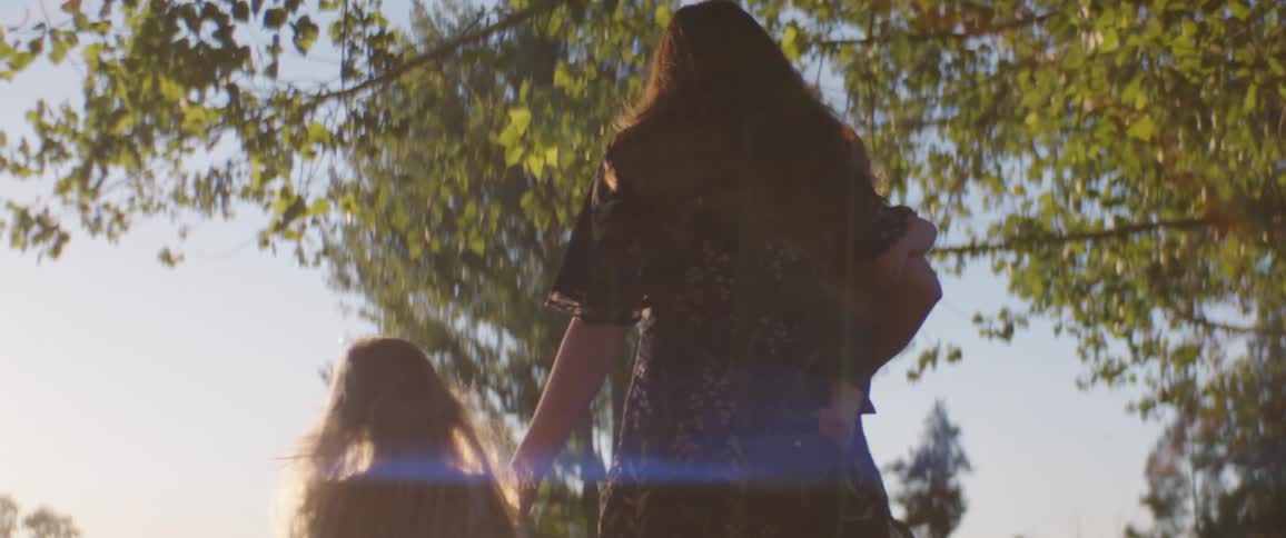 Mother walks past a tree and into a field while holding a baby in her arms and a child by her hand