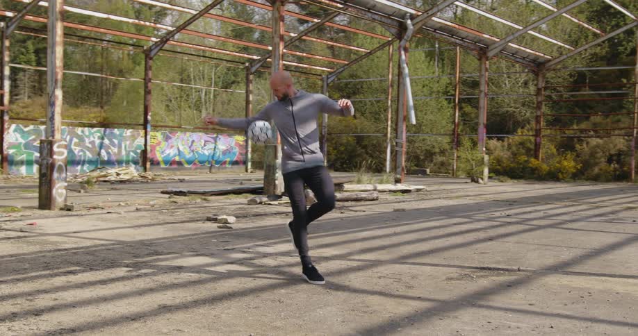 Man kicks a soccer ball up in the air inside a building frame near the woods
