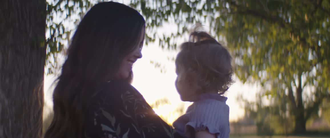 A mother smiles at her daughter by trees in a park as they smile