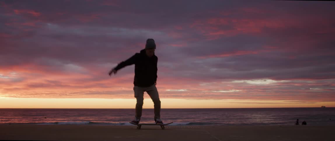 A man with a hat skateboarding on a boardwalk near the beach with the ocean the sky and clouds in the background