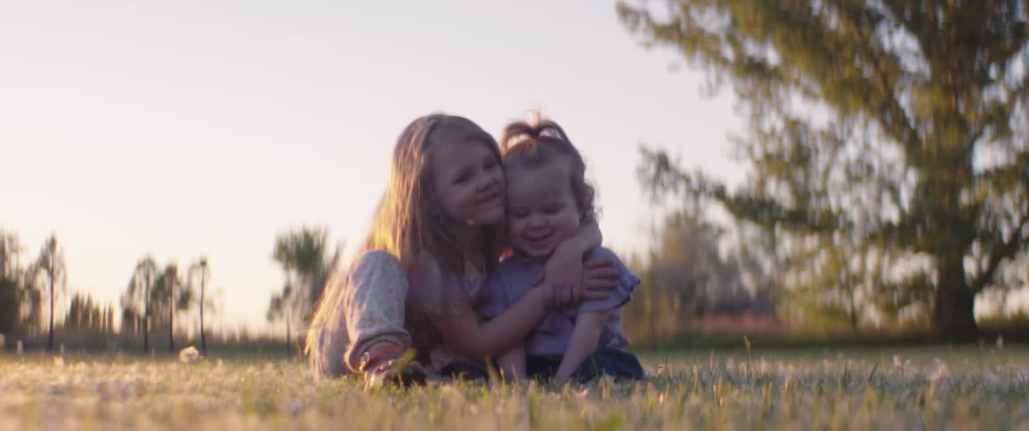 Female toddler sits on the grass with an infant and kisses the infant on the cheek