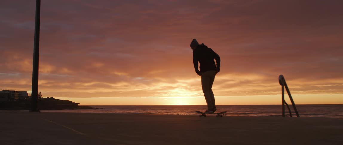 Skateboarder rides skateboard past staircase leading down toward beach silhouetted by sun sitting on horizon in red sky