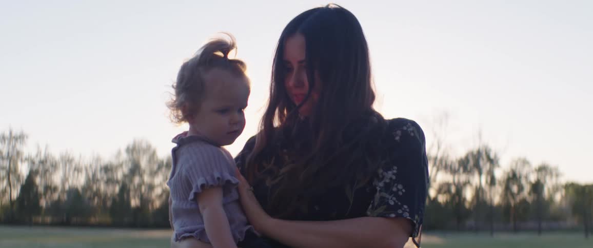A mother with long hair holds her baby and strokes her cheek while standing outdoors