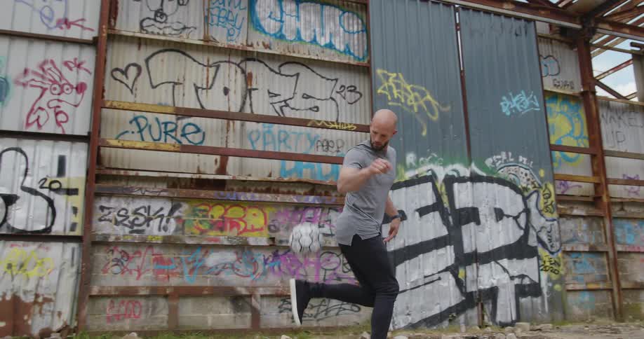 A male soccer player kicking a ball into the air in front of a graffitied wall