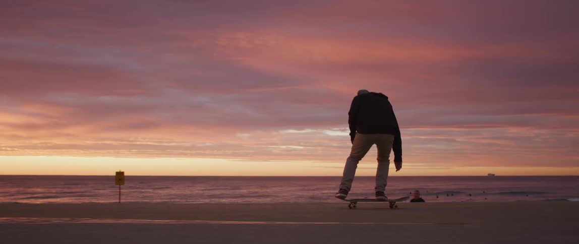 A skateboarder flips his board and jumps while skateboarding near the beach at sunset
