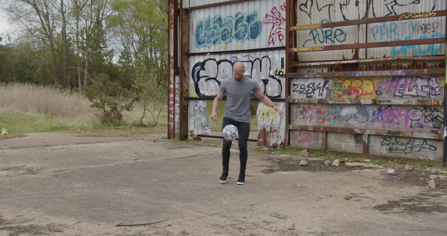 A male soccer player kicking a ball into the air in front of a graffitied wall