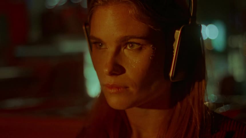 A woman wearing headphones and glitter on her face walks over to a booth and sits