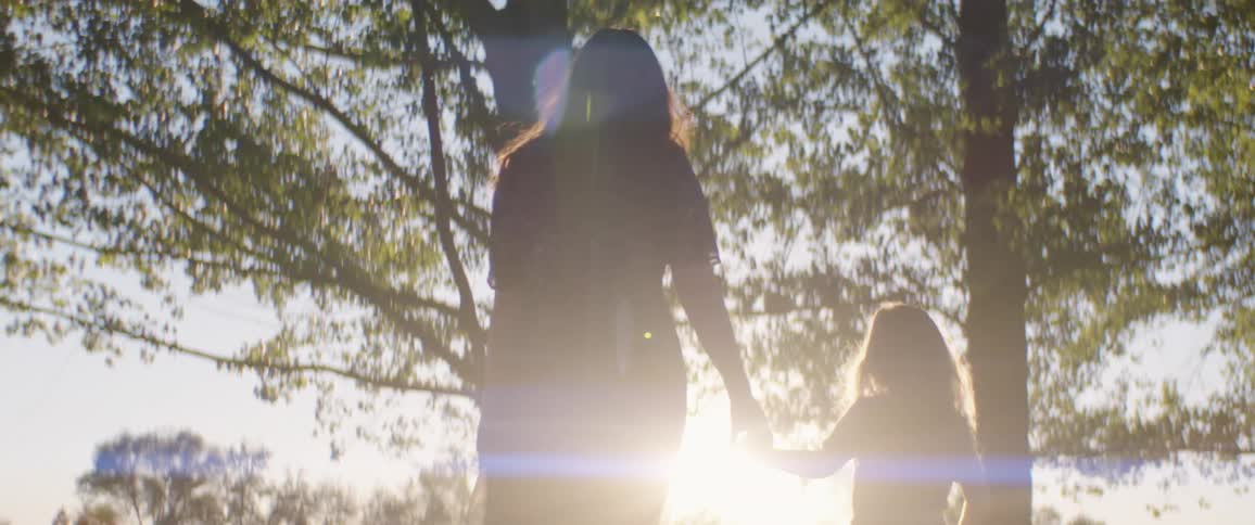 A mother and young daughter holding hands and walking through trees into the sunset