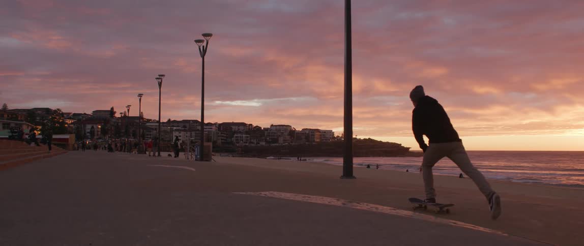 A man rides a skateboard along a walkway on a beach at sunset