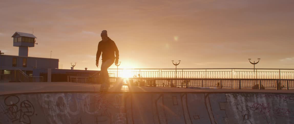 A skateboarder rides down a ramp in a park with the sun setting and shining on the horizon