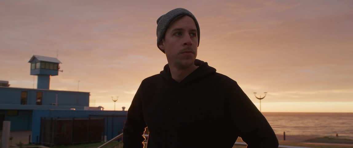 Man wearing a toque stands near an ocean and looks around as a woman walks a dog along the shoreline behind him