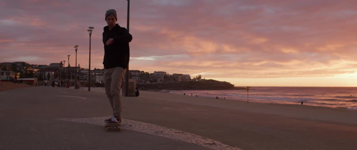 A man with a hat skateboarding on a boardwalk near the beach with the ocean the sky and clouds in the background