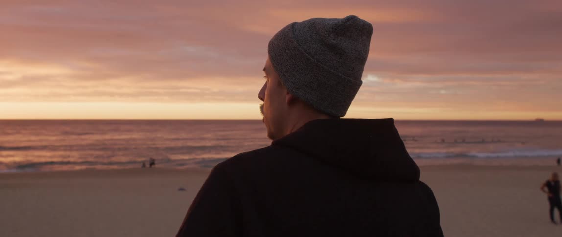 A man with a hat holding a skateboard on a boardwalk with the ocean sky and clouds in the background