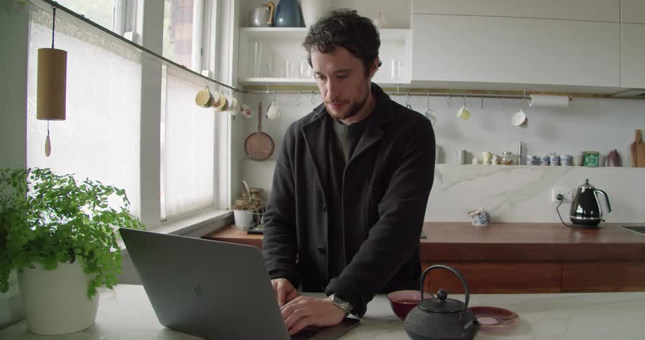 A man sips tea from a cup while typing on a laptop in a home kitchen during the day