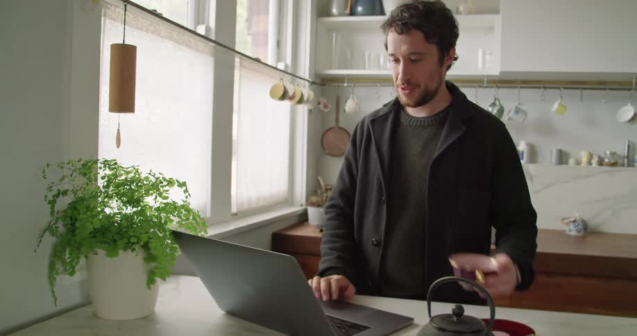 A man stands in his kitchen and drinks coffee while video chatting on his laptop