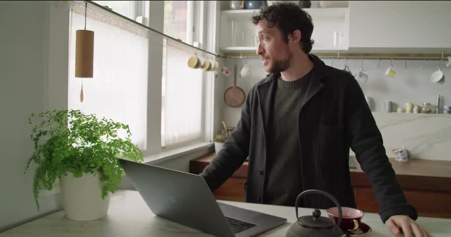 A man participates in a video chat using his laptop while standing in a kitchen during the day
