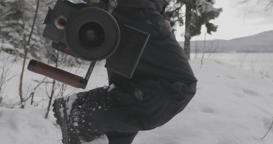 A man carries video equipment through a snow-covered area as he approaches a mountain lake