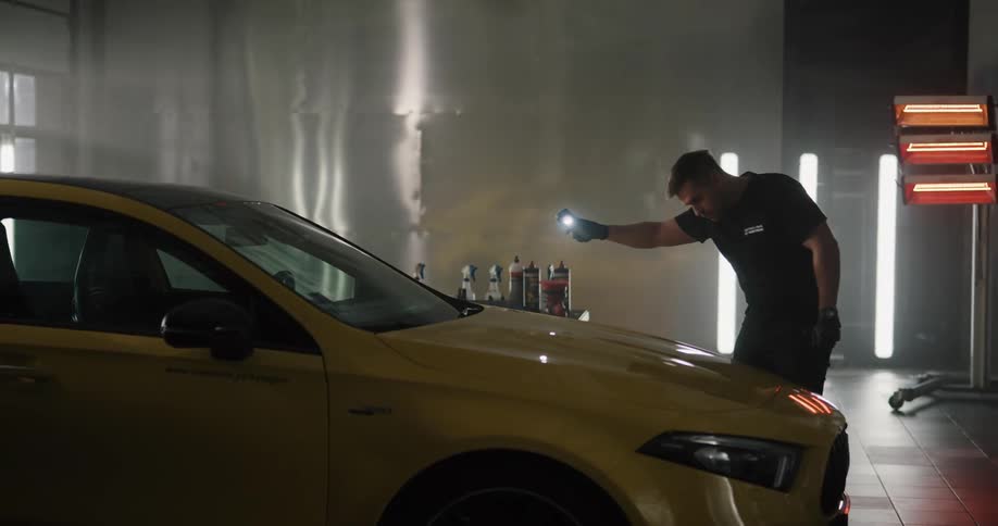 A man with gloves in the garage shining a flashlight on a vehicle