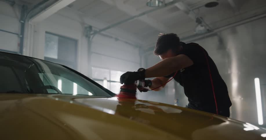 A man uses a powered car buffer to buff the hood of car in a garage