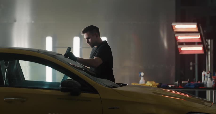 A man puts something from a bottle on a car windshield inside a commercial garage