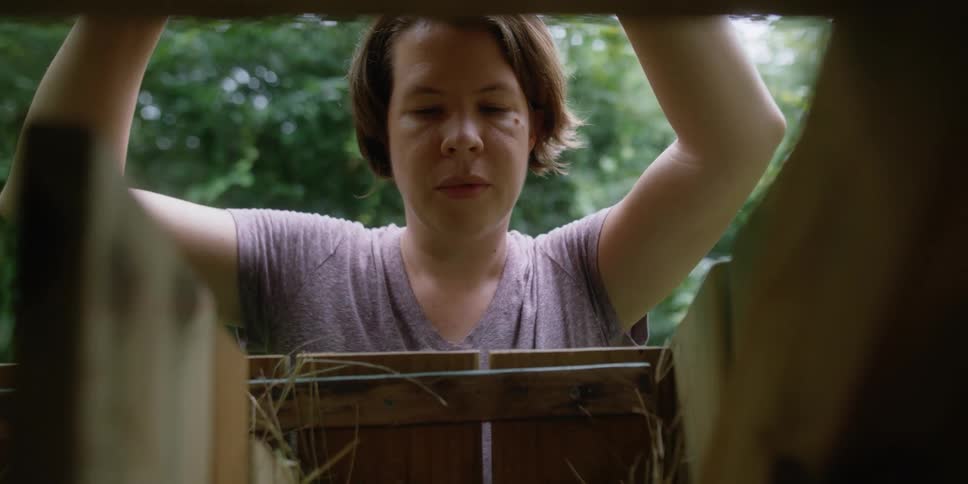 A woman opening a hatch and removing a egg from a chicken coop with trees the sky and clouds in the background