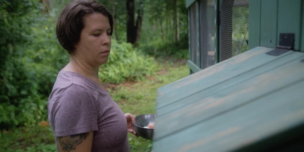 A woman closing a hatch on a chicken coop with trees the sky and clouds in the background