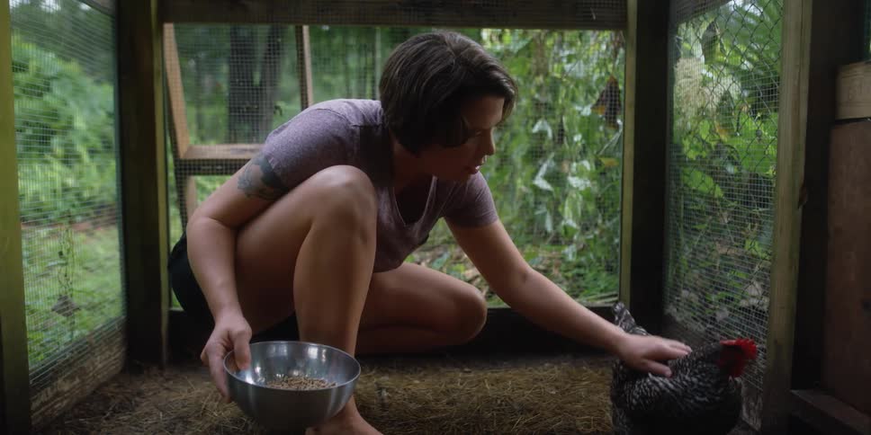 A woman inside a chicken coop pets a speckled hen while holding a bowl of food