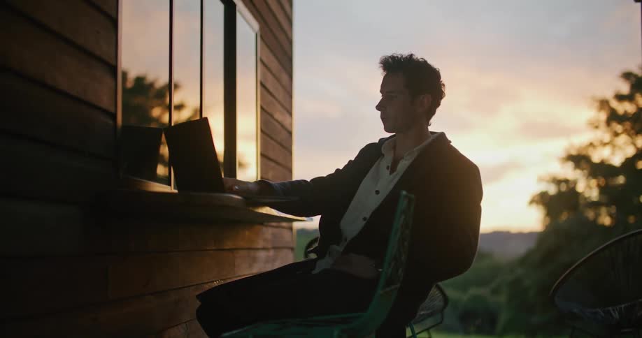 Man sits outdoors and types on a laptop that is sitting on a ledge attached to a building