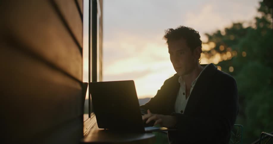 A man sitting on a deck working on a laptop sits back to think