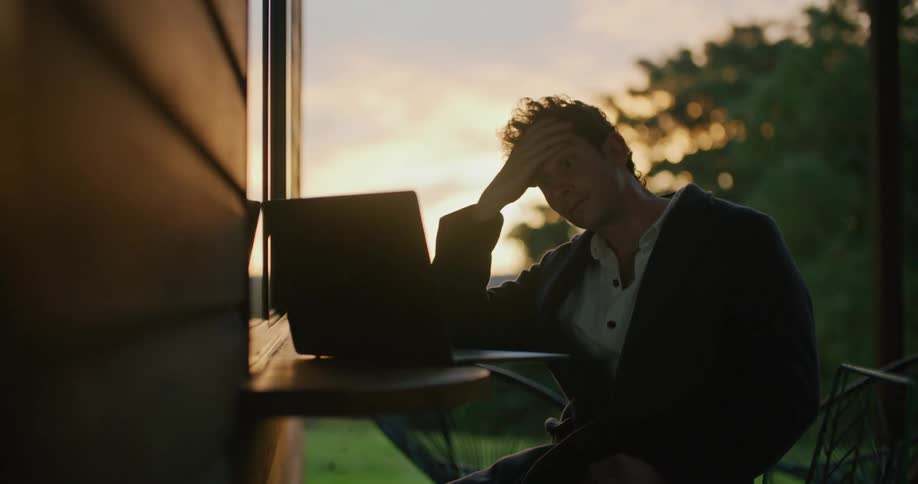 A man looks tired as he sits outside staring at his laptop computer
