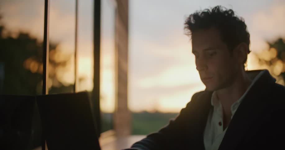 A man rubs his chin while thinking then types on his laptop while sitting on a deck at sunset