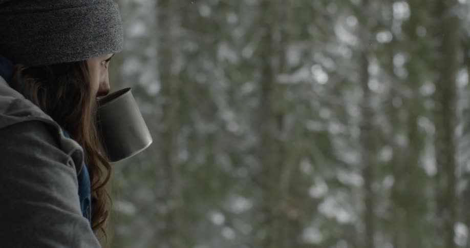 A woman takes a drink out of a mug looking out at the forest