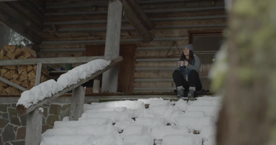 A woman sits on the snow-covered porch steps of a cabin drinking coffee