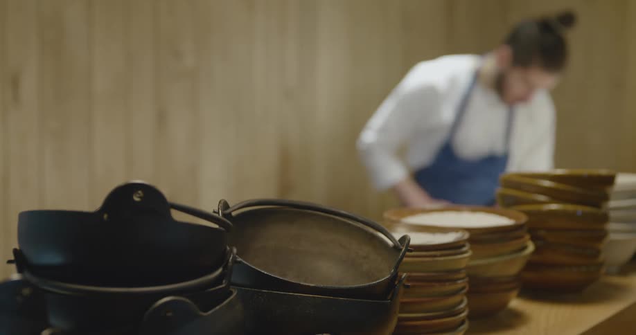 A cook in a restaurant works behind a counter covered in stacked pots and bowls