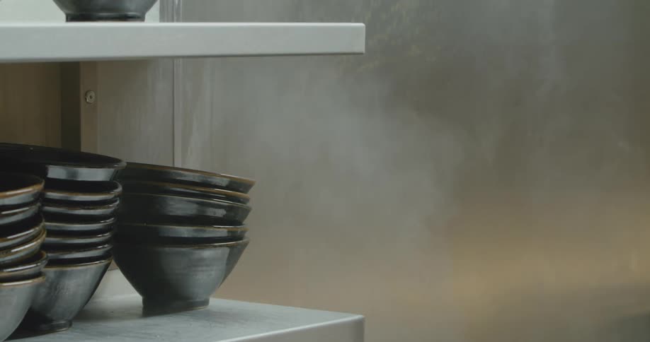 Steam rising from a vat of cooking noodles near bowls placed on a shelf