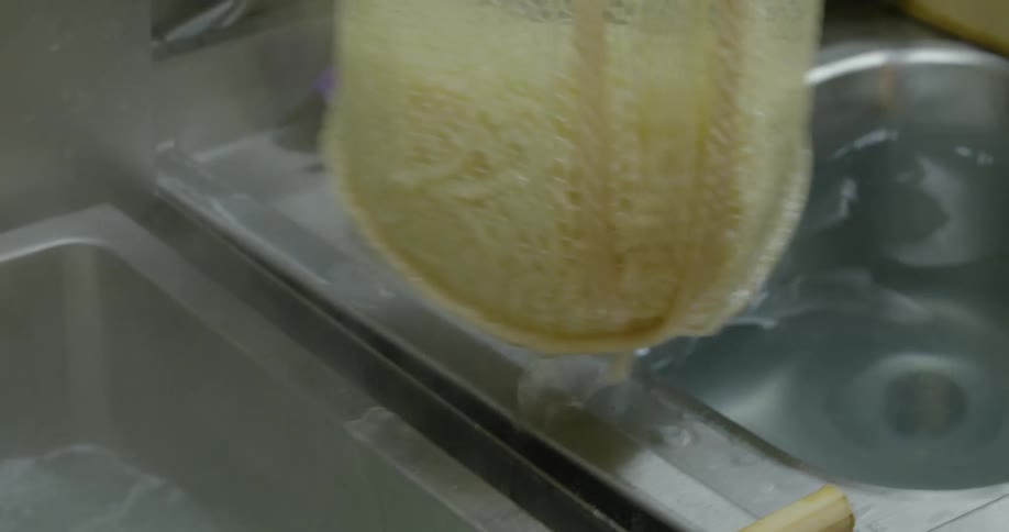 A cook uses a net to lift noodles from boiling water and dip them in a sink filled with cool water before spinning the noodles with their hands