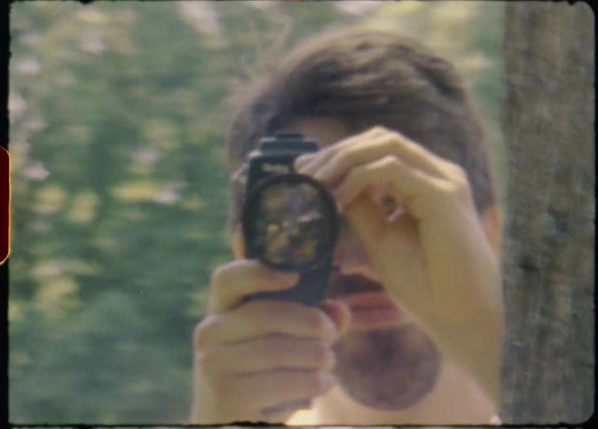 A man hides behind a tree while filming another person in a park