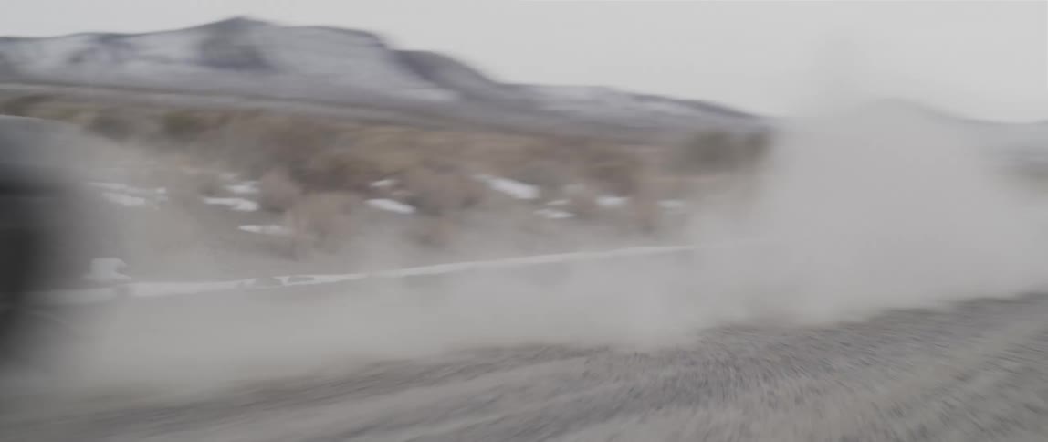 Sports car speeds along dirt road kicking up dust past snow dotted fields with snowy hills in background