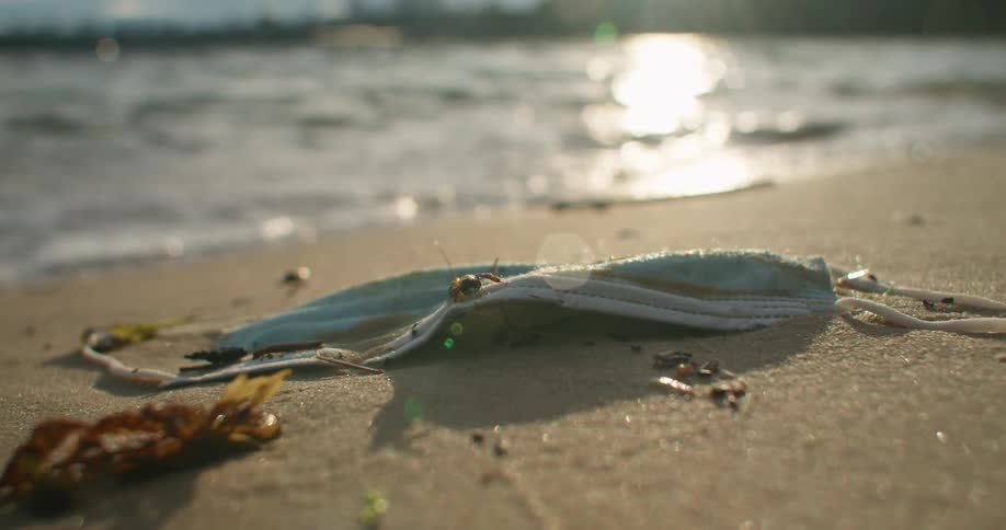 A discarded disposable face mask lays on the sand of a beach as waves wash ashore