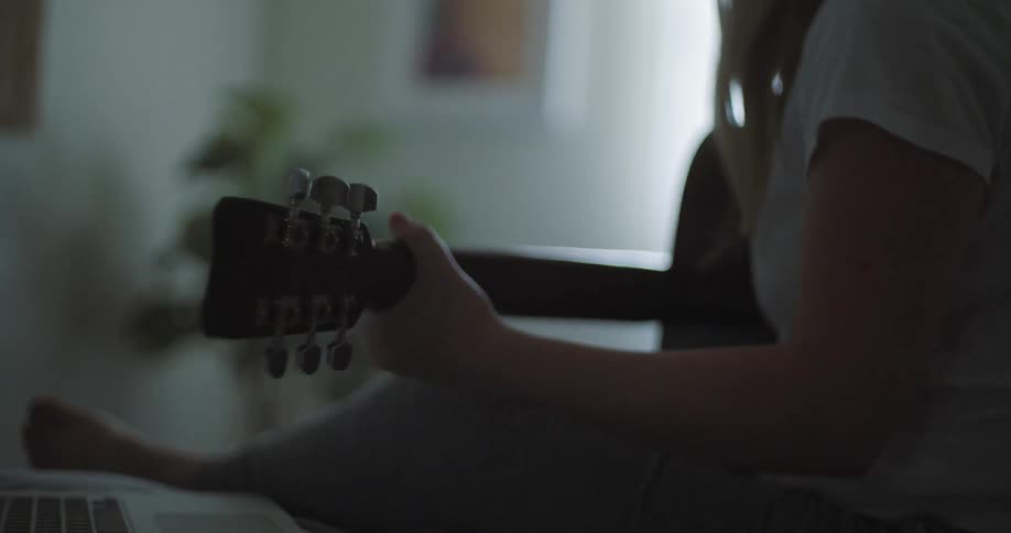 A young woman sits on her bed next to her laptop while practicing on her guitar