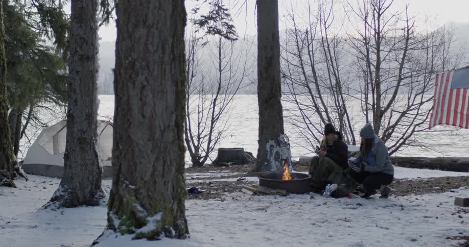 Two people sit down by a campfire near the water