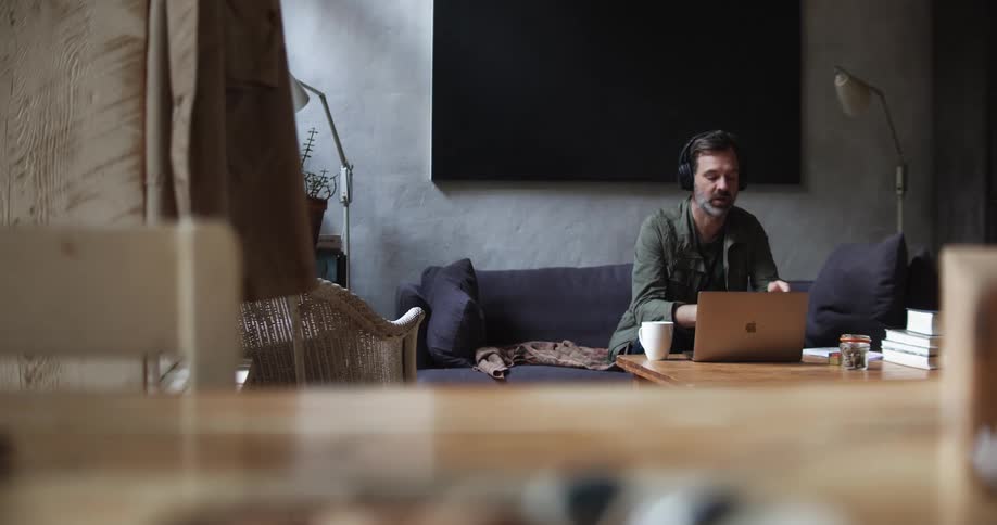 A man sits indoors on a couch while conversing with another person via his laptop