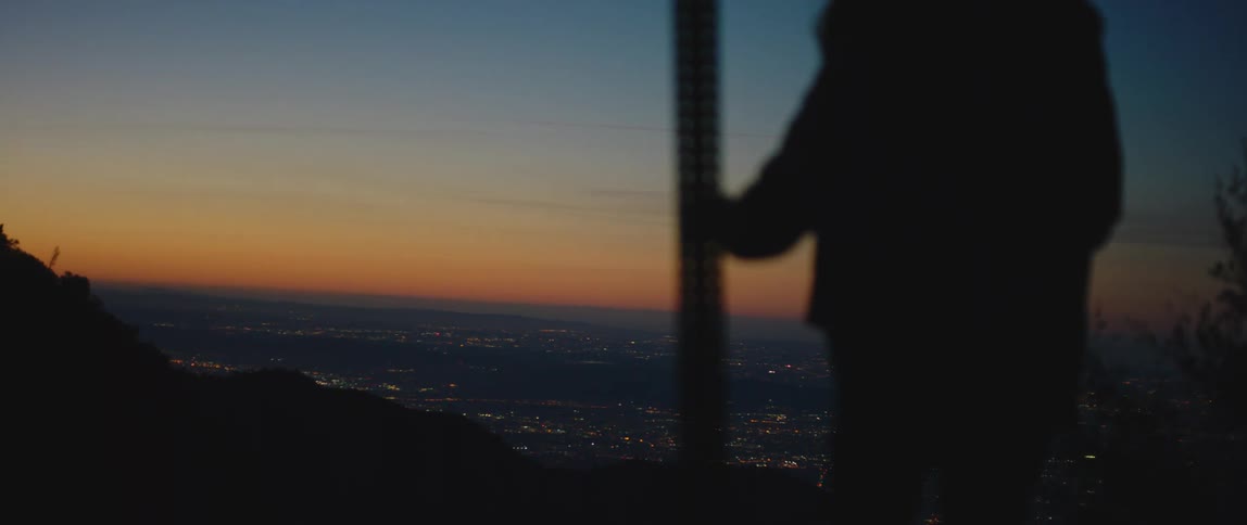 Woman stands on a hill and holds on to a post while looking out at the city at night