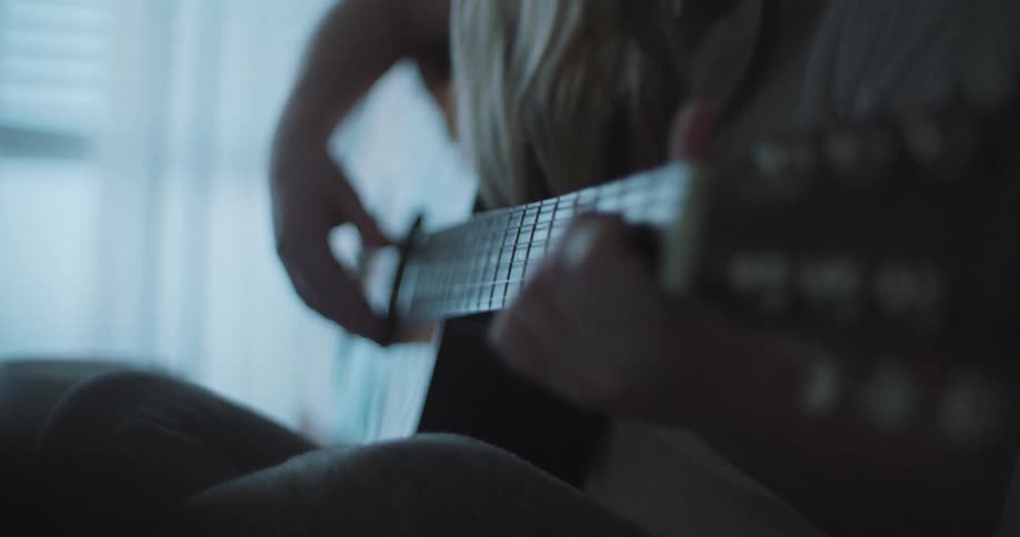 A young woman sits on her bed next to her laptop while practicing on her guitara young woman sits on her bed while strumming away on her guitar