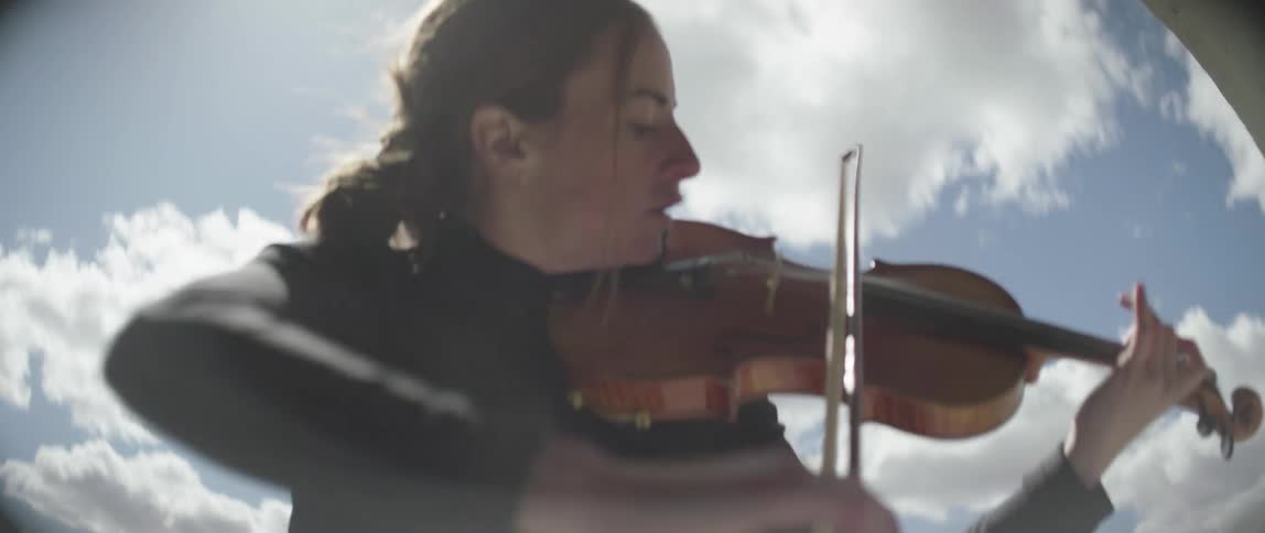 Woman plays string instrument energetically at mouth of concrete tunnel with mountains in distance