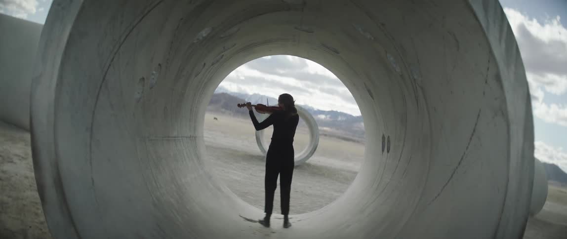 Female musician plays string instrument standing in section of concrete tunnel with mountains on horizon