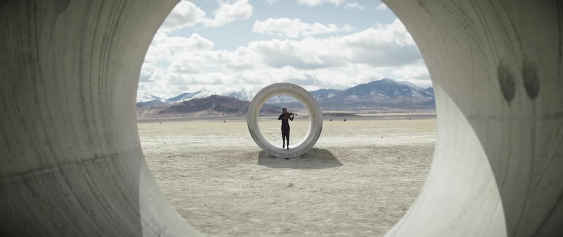 Silhouette of a female musician playing the violin and walking into a concrete pipe section with mountains behind