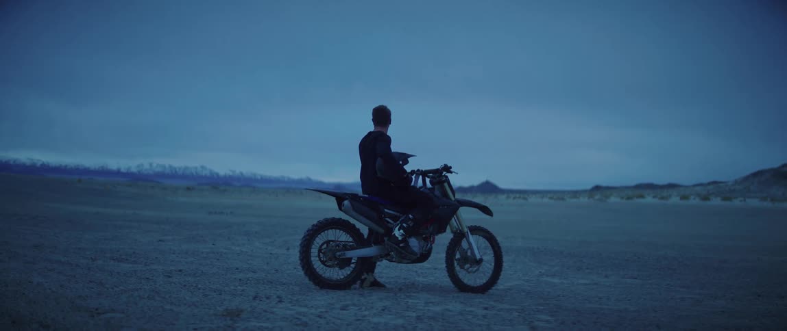 A man looks contemplatively into the distance while sitting atop an off road motorcycle parked in the desert on a cloudy evening