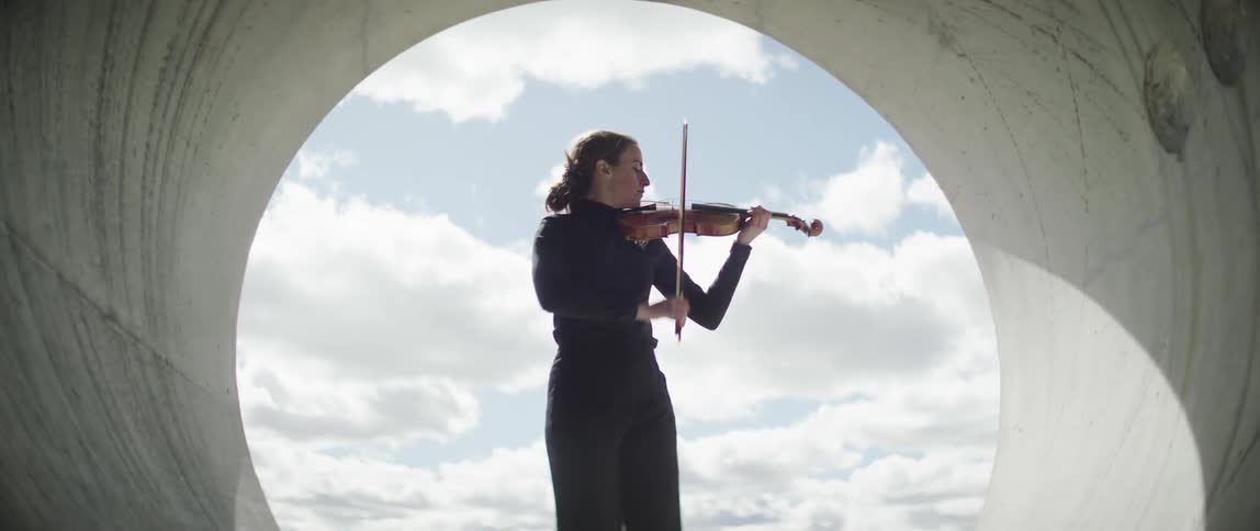 Woman plays violin near exit of large water tunnel under cloudy sky